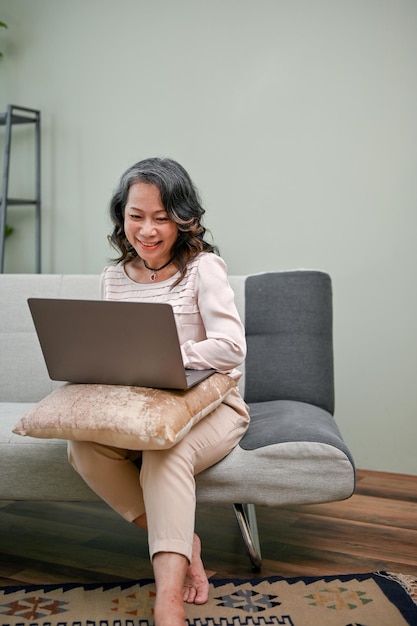 Gelukkige en vrolijke jaren '60 verouderde vrouw die laptop gebruikt terwijl ze op haar bank zit