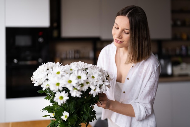 Foto gelukkige en vrolijke jonge vrouw in het wit die witte bloemen schikt thuis in de keuken