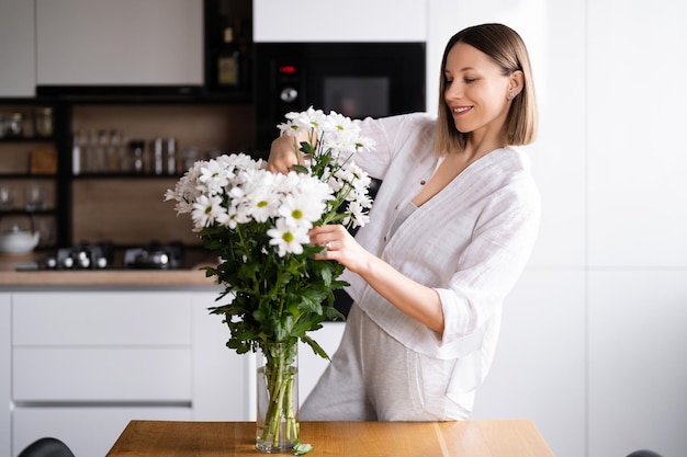 Gelukkige en vrolijke jonge vrouw in het wit die witte bloemen schikt thuis in de keuken