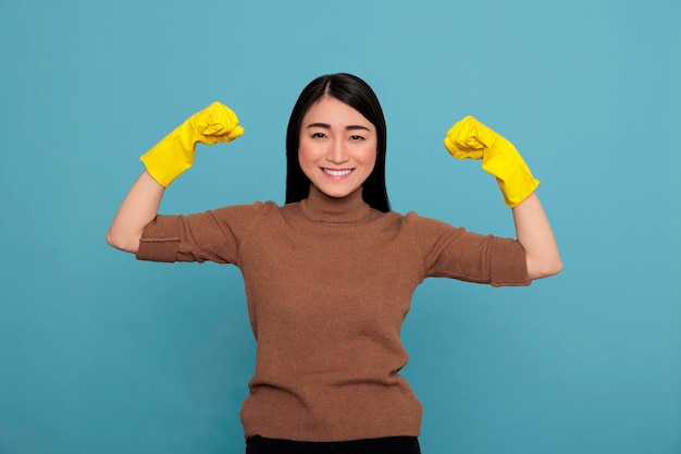 Gelukkige energieke mooie Aziatische huisvrouw die armen opheft en biceps geïsoleerde blauwe achtergrond laat zien, huisvrouw werknemer, schoonmaak thuis concept, vrouw glimlacht na het voltooien van zijn baan,