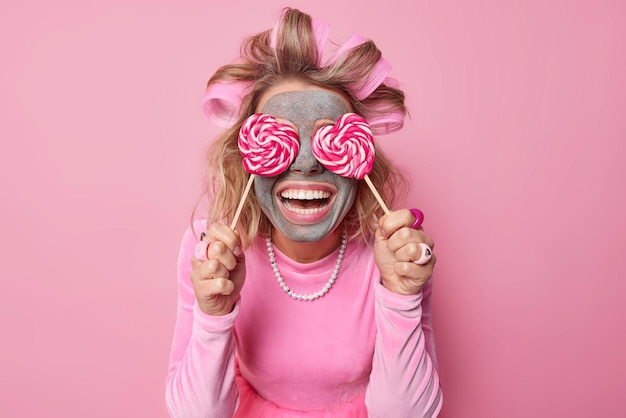 Foto gelukkige europese vrouw bedekt ogen met karamelsuikergoed glimlacht graag heeft zoetekauw draagt haarrollen en jurk ketting past kleimasker toe voor huidbehandeling geïsoleerd over roze achtergrond