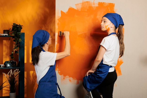 Gelukkige familie die plezier heeft met het schilderen van muren met oranje verf, de kleur van het oppervlak verandert met renovatiegereedschap, ladder en penseel. Volwassen en klein kind met borstel met borstelharen en roller.