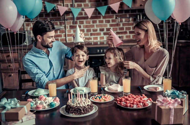 Gelukkige familie heeft plezier terwijl ze aan tafel zitten in de ingerichte keuken tijdens het verjaardagsfeest, ouders zetten verjaardagshoedjes op hun kinderen