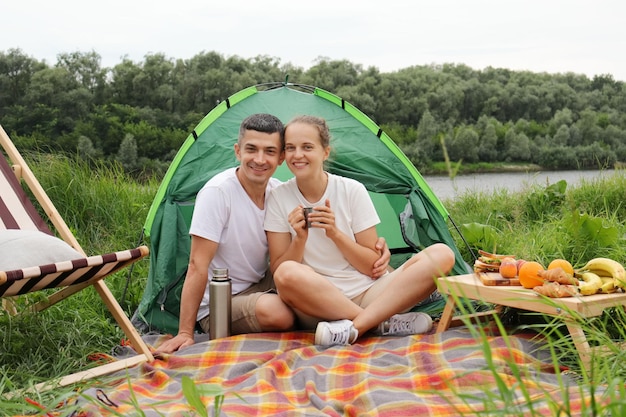 Gelukkige familie in het weekend vrouw en man ontspannen in de buurt van de rivier met een picknick genietend van koffie of thee kijkend naar de camera met een glimlach