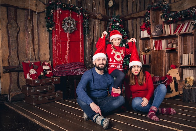 Gelukkige familie in kerstmutsen zittend in een kamer in een rustieke stijl Dochter heft hoeden op aan ouders