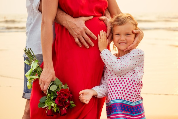 Gelukkige familie jonge man, mooie zwangere vrouw in rode jurk met een boeket rozen en hun schattige dochtertje knuffelen zee romantische zonsondergang zomer. De relatie tussen ouders en kinderen.