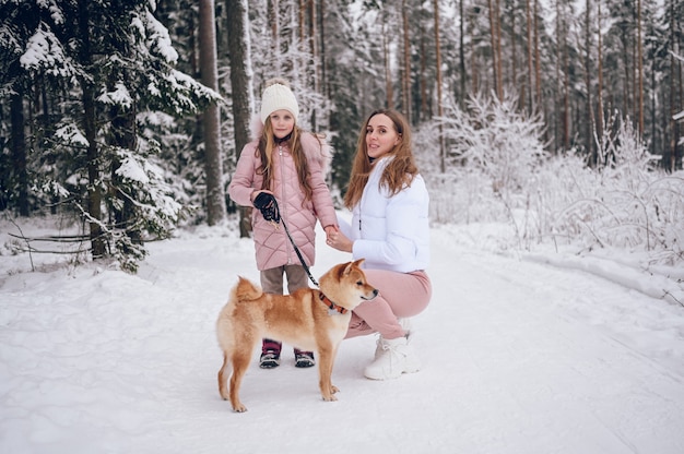 Gelukkige familie jonge moeder en schattig meisje in roze warme uitloper wandelen met plezier met rode shiba inu hond in besneeuwde witte koude winter forest buitenshuis