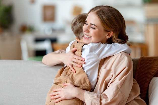 Gelukkige familie jonge vrouw en kleine jongen in casual kleding omhelzen en zoenen terwijl feliciteren met vakantie moederdag in zonnige kamer thuis