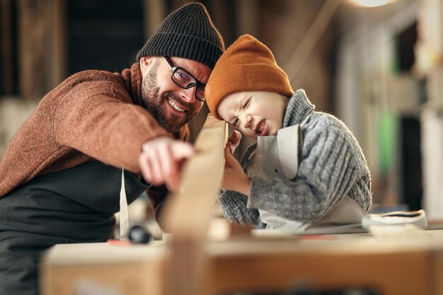 Foto gelukkige familie kleine jongen en vader in truien en hoeden die het bord bij elkaar houden tijdens het werken met hout in een moderne handwerkstudio