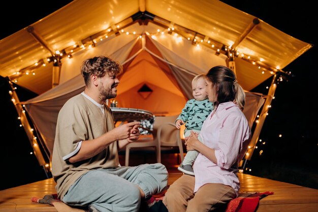 Gelukkige familie met mooie baby ontspannen en samen tijd doorbrengen in glamping op zomeravond in de buurt van gezellig vreugdevuur Luxe kampeertent voor openluchtrecreatie en recreatie Lifestyle-concept