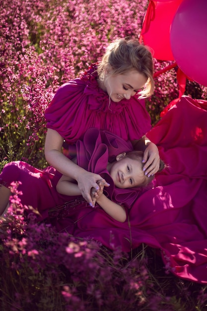 Gelukkige familie moeder en dochter in roze jurken zitten in een veld met bloemen en grote ballonnen bij zonsondergang in de zomer