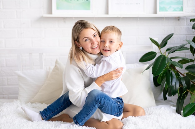 Gelukkige familie moeder en zoon in witte truien en jeans op het bed in een lichte slaapkamer thuis knuffelen en glimlachen