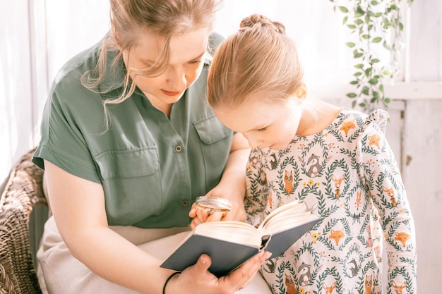 Gelukkige familie moeder met schattig klein kind dochter meisje veel plezier samen in buitenwijk platteland huis op zonnige zomerdag openhartige moeder knuffelen en leest boek aan kind verkennen boek met vergrootglas