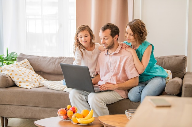 Gelukkige familie, moeder, vader en dochter zitten op de bank en kijken samen naar een laptopmonitor.