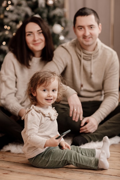 Gelukkige familie, moeder, vader en dochtertje spelen met mobiel en zitten in de buurt van de kerstboom thuis.