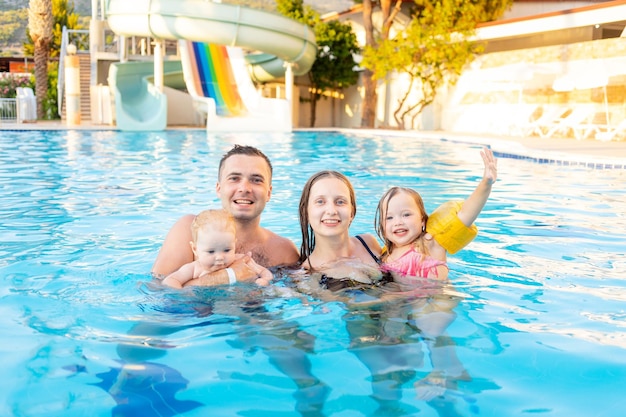 Gelukkige familie, moeder, vader en twee kinderen zwemmen in het zwembad met glijbanen en hebben plezier op vakantie, glimlachen en lachen