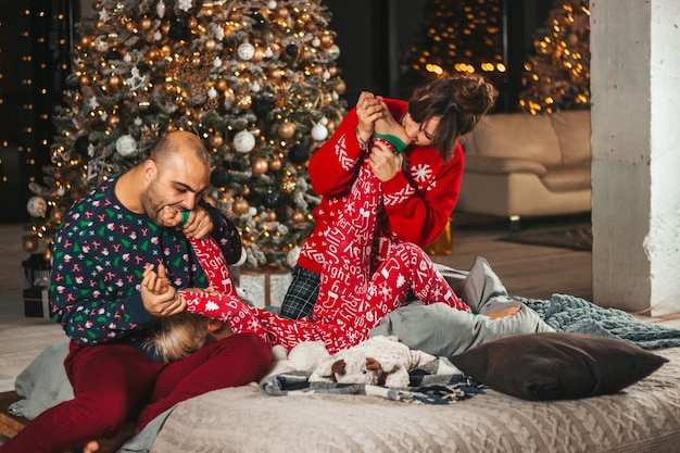 Gelukkige familie op het bed in de buurt van de kerstboom spelen met het kleine kind bijtplezier