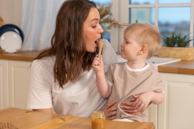 Gelukkige familie thuis, moeder die baby voedt in de keuken, kleine jongen met een rommelig, grappig gezicht, eet gezond fo