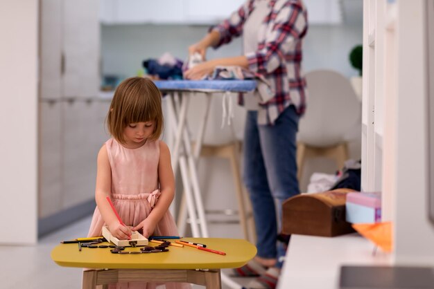 Gelukkige familie tijd samen doorbrengen thuis schattig dochtertje in een roze jurk spelen en schilderen van de juwelendoos terwijl jonge roodharige moeder kleren achter haar strijkt