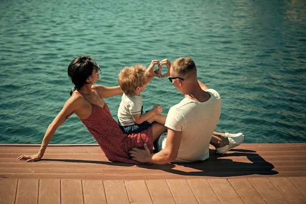 Gelukkige familie tijd samen doorbrengen, zee achtergrond. Ouders met zoon zitten aan de kust, achteraanzicht. Moeder en vader maken hart- of liefdegebaar met handen in de buurt van hun kind. Familie vakantie concept.