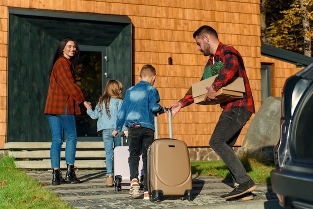 Gelukkige familie van ouders en twee kinderen die koffers van de auto naar het nieuwe huis dragen. Concept van verhuizen.
