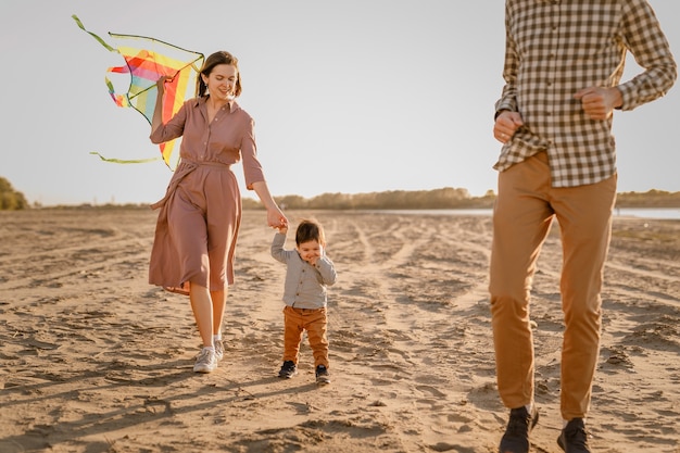 Gelukkige familie wandelen op het zandstrand van de rivier. vader, moeder die zoontje op handen houdt en met vlieger speelt.