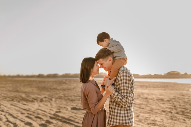 Gelukkige familie wandelen op het zandstrand van de rivier. Vader, moeder die zoontje op handen houdt en samen gaat. Achteraanzicht. Familiebanden concept.