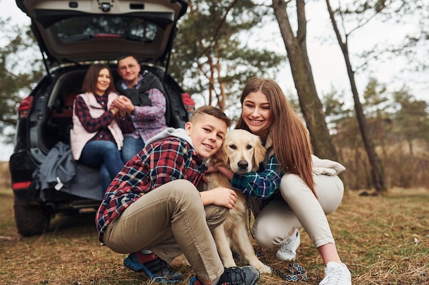 Gelukkige familie zitten en plezier hebben met hun hond in de buurt van moderne auto buiten in het bos