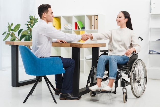 Foto gelukkige gehandicapte jonge vrouwenzitting op rolstoel het schudden handen met mannelijke collega in het bureau