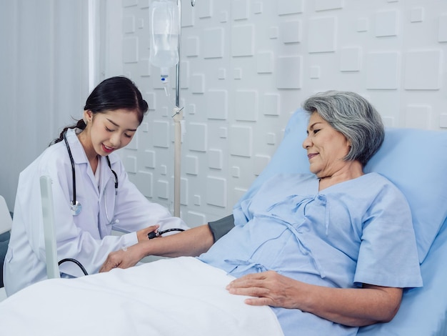 Gelukkige glimlach Aziatische oudere oude vrouwelijke patiënt in lichtblauwe jurk liggend op bed terwijl mooie jonge vrouw arts in wit pak bloeddruk meten en pols controleren in ziekenhuiskamer