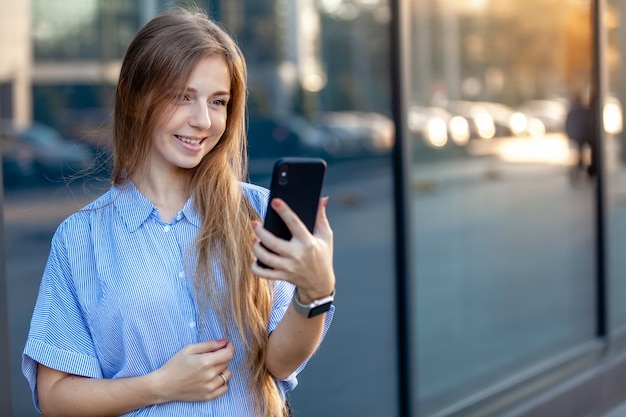 Gelukkige glimlachende aantrekkelijke jonge vrouw die een smartphone gebruiken dichtbij het bureaugebouw