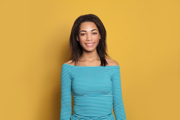 Gelukkige glimlachende gemengde ras etnisiteit zwarte jonge vrouw in blauw gestreepte hemd op heldergele studio achtergrond