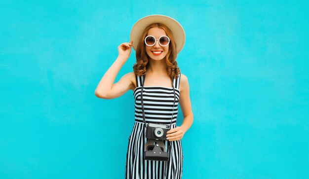 Gelukkige glimlachende jonge vrouwelijke fotograaf met filmcamera in zomer strohoed op blauwe achtergrond