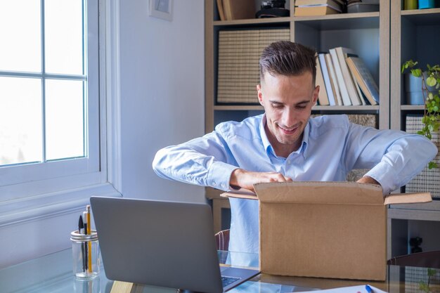 Foto gelukkige glimlachende man die werkt en een kleine doos opent die net thuis is afgeleverd en geniet van zijn verrassing alleen plezier te hebben jonge blanke man die een cadeautje of geschenk op kantoor opdoet