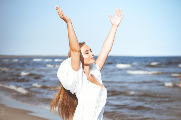Gelukkige glimlachende mooie vrouw op het oceaanstrand die in een witte zomerjurk staat en handen opheft.