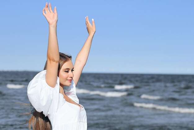 Gelukkige glimlachende mooie vrouw op het oceaanstrand die in een witte zomerjurk staat en handen opheft.