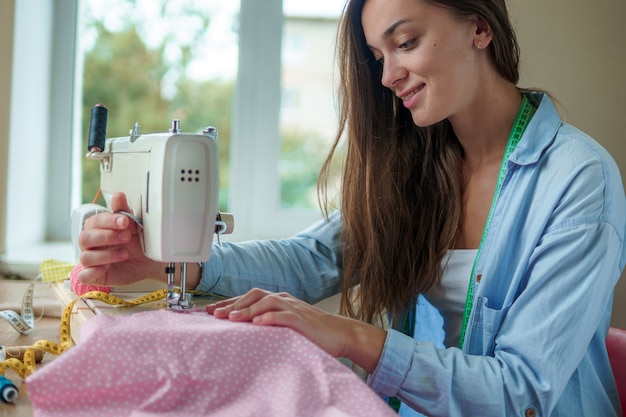 Gelukkige glimlachende naaister met elektrische naaimachine en verschillende naaiende toebehoren voor het naaien van kleren op het werk