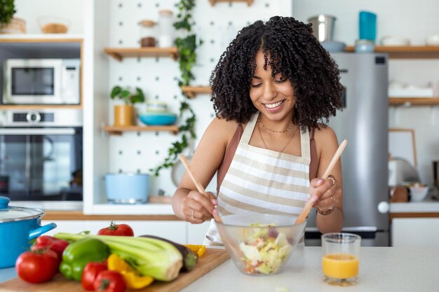 Foto gelukkige glimlachende schattige vrouw bereidt een verse gezonde veganistische salade met veel groenten in de keuken thuis en probeert een nieuw recept