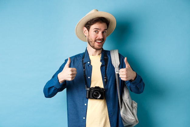 Gelukkige glimlachende toerist zegt ja en duimen opdagen, op zomervakantie gaan, rugzak en fotocamera, blauwe achtergrond vasthouden.