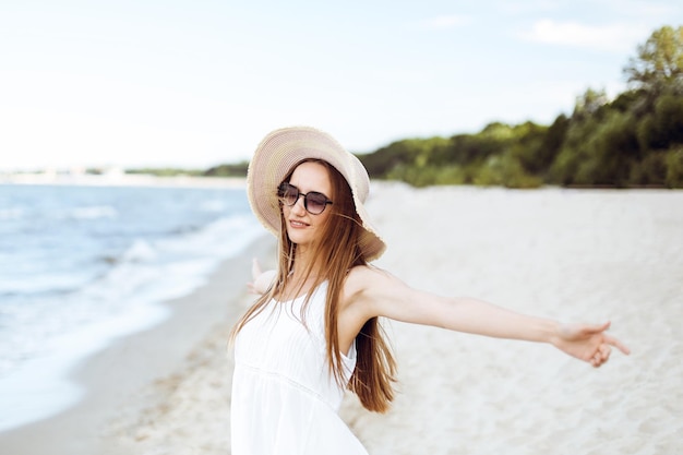 Gelukkige glimlachende vrouw in vrij gelukzaligheid op oceaanstrand die zich met een hoed, zonnebril, en open handen bevindt. Portret van een multicultureel vrouwelijk model in witte zomerjurk genietend van de natuur tijdens het reizen