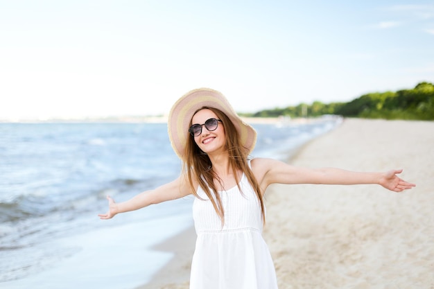 Gelukkige glimlachende vrouw in vrij gelukzaligheid op oceaanstrand die zich met een hoed, zonnebril, en open handen bevindt. Portret van een multicultureel vrouwelijk model in witte zomerjurk genietend van de natuur tijdens het reizen