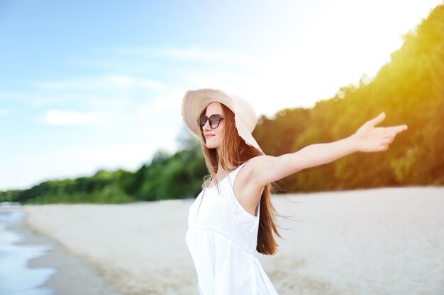 Gelukkige glimlachende vrouw in vrij gelukzaligheid op oceaanstrand die zich met een hoed, zonnebril, en open handen bevindt. Portret van een multicultureel vrouwelijk model in witte zomerjurk genietend van de natuur