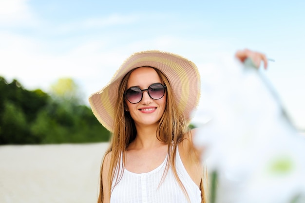 Gelukkige glimlachende vrouw in vrij gelukzaligheid op oceaanstrand die zich met hoed, zonnebril, en witte bloemen bevindt. Portret van een multicultureel vrouwelijk model in witte zomerjurk genietend van de natuur tijdens het reizen