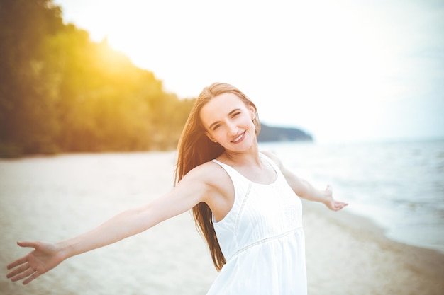 Gelukkige glimlachende vrouw in vrije gelukzaligheid op het oceaanstrand met open handen. Portret van een multiculturele vrouwelijk model in witte zomerjurk die geniet van de natuur tijdens reisvakantie vakantie