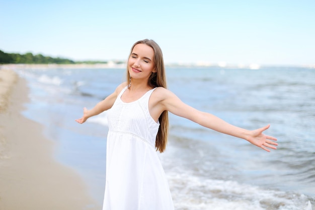 Gelukkige glimlachende vrouw in vrije gelukzaligheid op het oceaanstrand staande met open handen. Portret van een multicultureel vrouwelijk model in witte zomerjurk die geniet van de natuur tijdens reizen vakanties vakantie outd