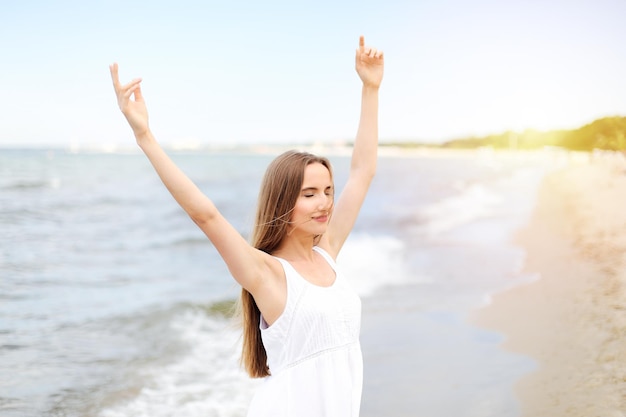 Gelukkige glimlachende vrouw in vrije gelukzaligheid op oceaanstrand dat zich met het opheffen van handen bevindt. Portret van een multicultureel vrouwelijk model in witte zomerjurk genietend van de natuur