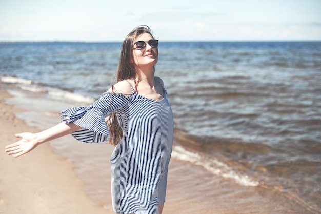 Gelukkige glimlachende vrouw in vrije gelukzaligheid op oceaanstrand dat zich met open handen bevindt. Portret van een brunette vrouwelijk model in zomerjurk genietend van de natuur tijdens reisvakanties vakantie buitenshuis.