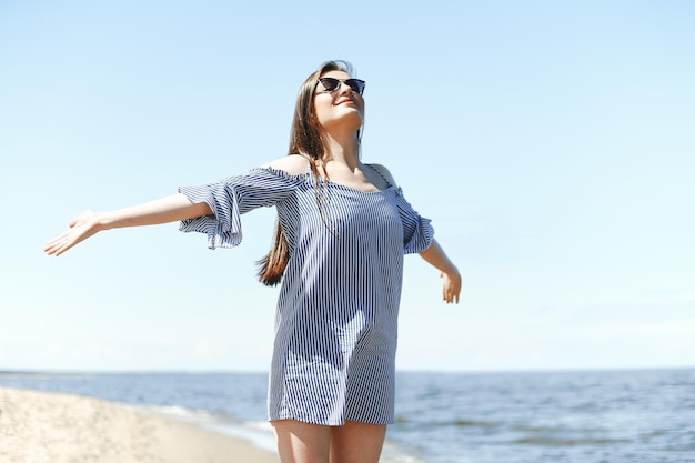 Gelukkige glimlachende vrouw in vrije gelukzaligheid op oceaanstrand dat zich met open handen bevindt. Portret van een brunette vrouwelijk model in zomerjurk genietend van de natuur tijdens vakantie vakantie buitenshuis