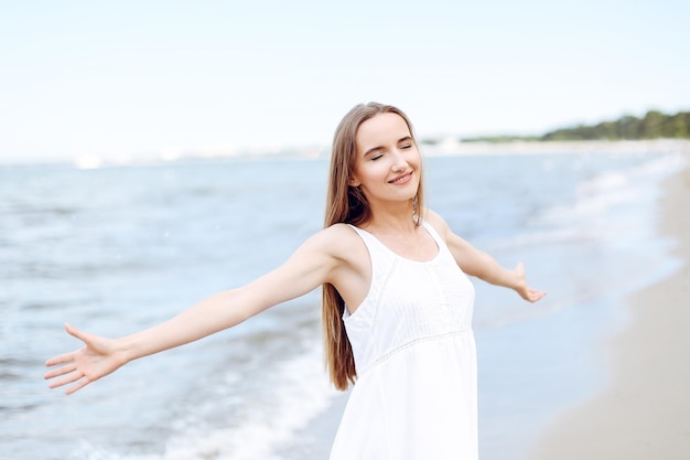 Gelukkige glimlachende vrouw in vrije gelukzaligheid op oceaanstrand dat zich met open handen bevindt. Portret van een multicultureel vrouwelijk model in een witte zomerjurk die geniet van de natuur tijdens vakanties
