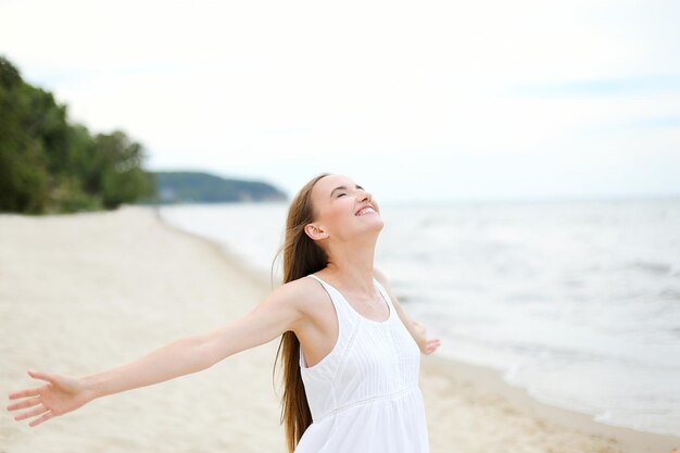 Gelukkige glimlachende vrouw in vrije gelukzaligheid op oceaanstrand dat zich met open handen bevindt. Portret van een multicultureel vrouwelijk model in een witte zomerjurk die geniet van de natuur tijdens vakanties